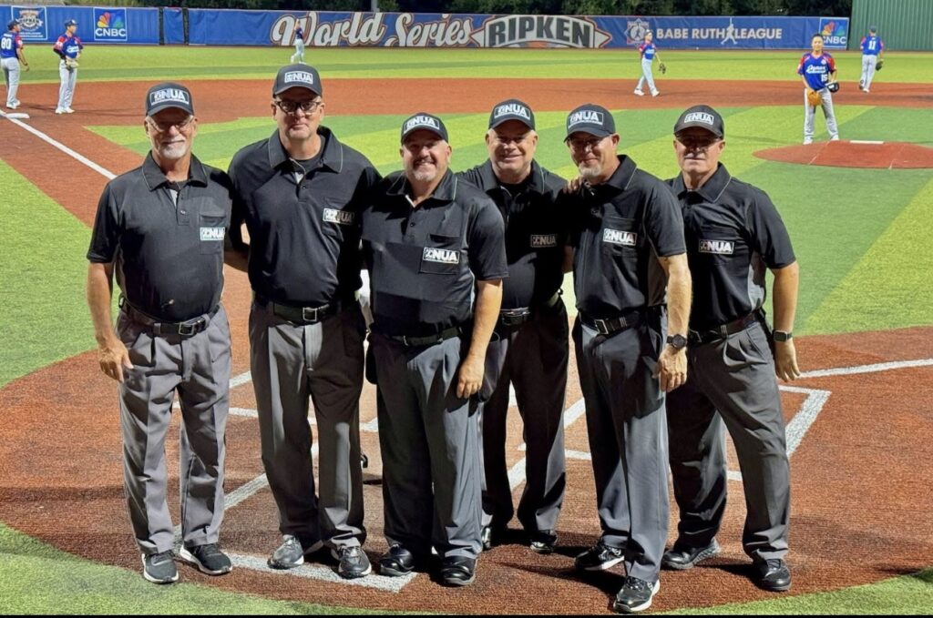 Group of six baseball umpires in black uniforms standing together on the field at a World Series youth baseball game.